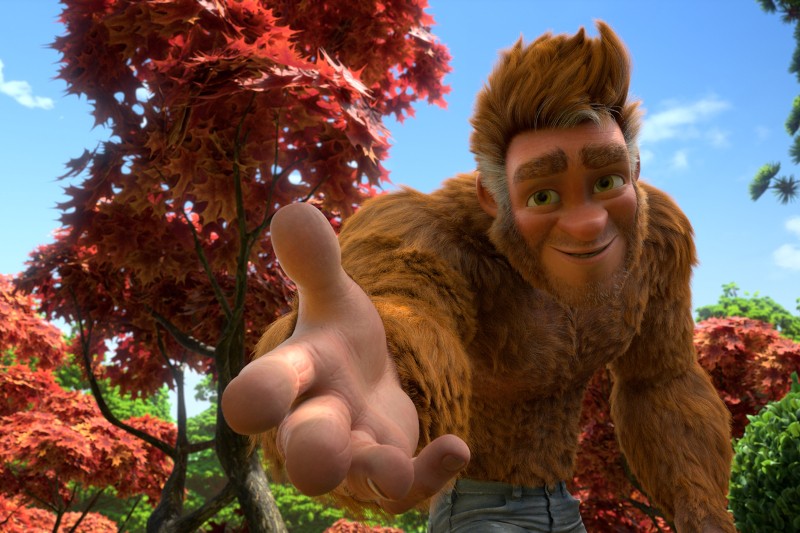 Przyjazna, animowana, brązowo-futrzana postać o ludzkich cechach uśmiecha się i wyciąga rękę do przodu. Żywe, czerwonolistne drzewa i zielone liście wypełniają słoneczne tło.