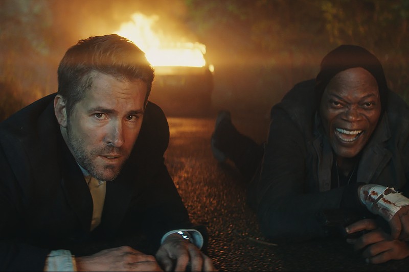 Dwóch mężczyzn leży nocą na mokrej drodze, jeden wygląda poważnie, a drugi się śmieje. Za nimi widać płonący samochód, a w tle drzewa.