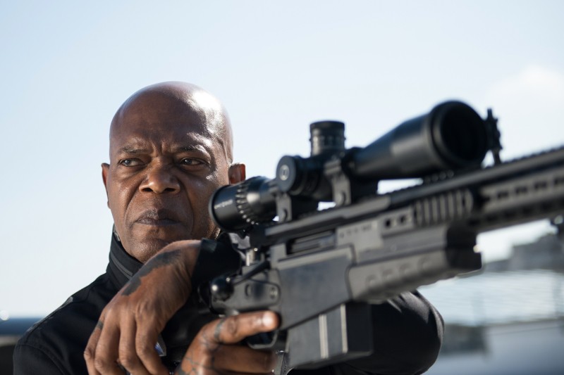 Poważnie wyglądający mężczyzna ubrany na czarno celuje z dużego karabinu snajperskiego z lunetą, na zewnątrz, w jasny dzień.