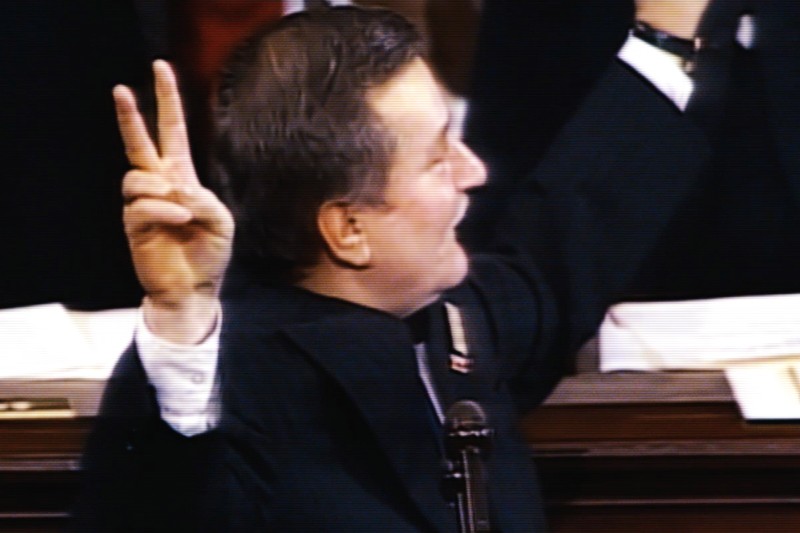 Mężczyzna w ciemnym garniturze uśmiecha się i podnosi rękę, wykonując gest pokoju z dwoma uniesionymi palcami. Stoi na podium, a w tle widać rozmazanych ludzi.