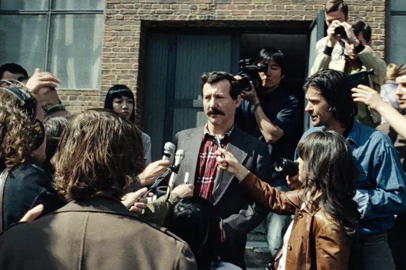 Mężczyzna z wąsami stoi przed ceglanym budynkiem, otoczony przez reporterów trzymających mikrofony i kamery, którzy przeprowadzają z nim wywiad w zatłoczonej, pełnej napięcia scenie.