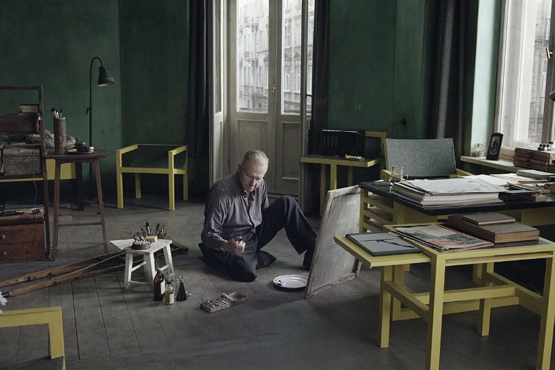 Starszy mężczyzna siedzi na podłodze w ciemnym pokoju o zielonych ścianach, malując na płótnie. Otaczają go przybory artystyczne i żółte meble, a duże okna wpuszczają miękkie światło dzienne.