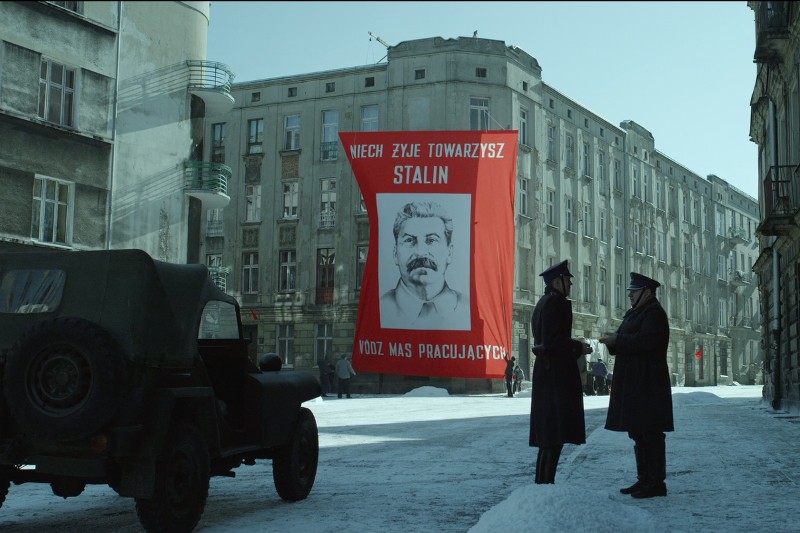 Dwóch umundurowanych mężczyzn rozmawia na zaśnieżonej ulicy przed dużym czerwono-białym banerem z portretem Stalina i polskim napisem, w otoczeniu starych budynków mieszkalnych i pojazdu wojskowego.