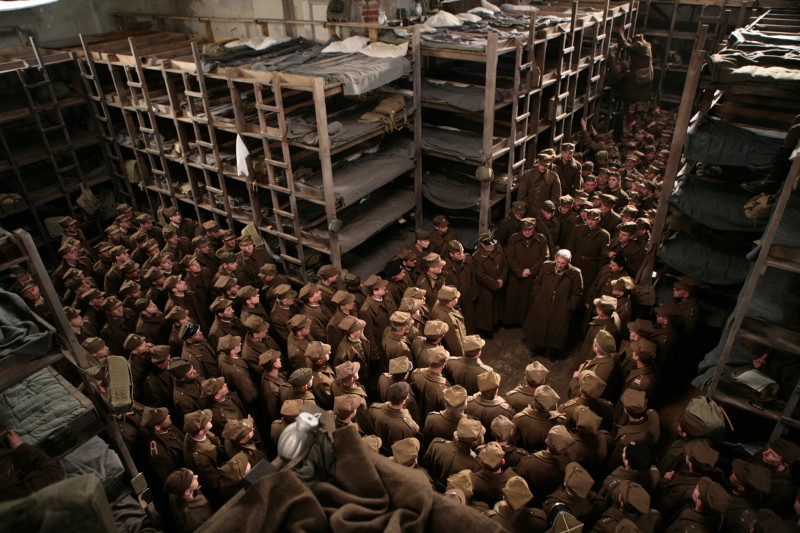Duża grupa żołnierzy w mundurach stoi blisko siebie w wąskim przejściu między wysokimi, zatłoczonymi łóżkami piętrowymi w słabo oświetlonym koszarze lub dormitorium. Atmosfera wydaje się napięta i poważna.