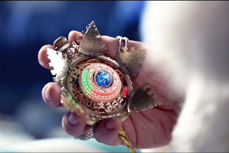 Dłoń trzyma złoty, ozdobny, okrągły amulet ze skomplikowanymi wzorami, świecącymi kolorowymi światłami i trójkątnymi punktami na krawędziach. Obiekt wydaje się mistyczny i złożony.