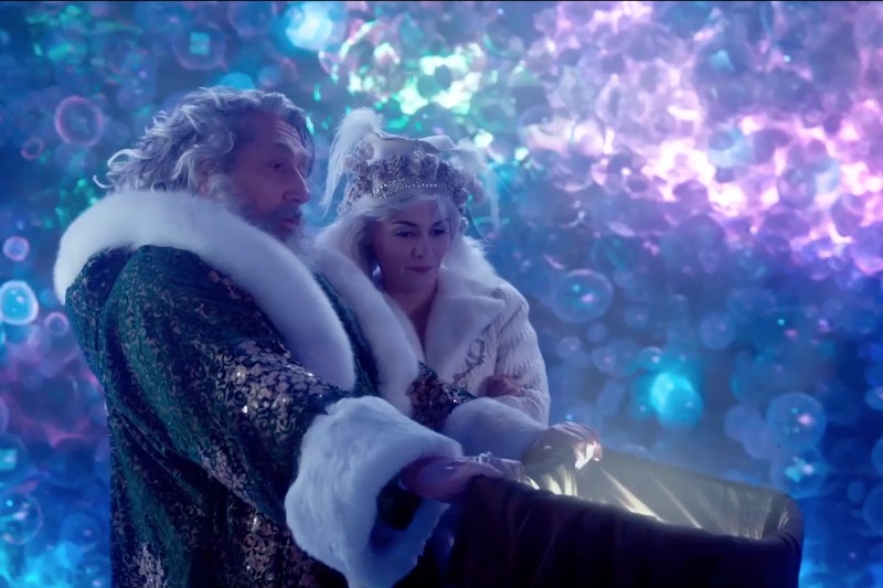 Starszy mężczyzna i kobieta w ozdobnych, podszytych futrem strojach stoją razem i patrzą na błyszczącą, otwartą klatkę piersiową na tle żywego, kolorowego, przypominającego bańkę tła.