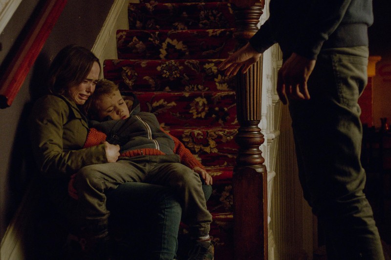 Zrozpaczona osoba siedzi na schodach, trzymając śpiące lub nieprzytomne dziecko, podczas gdy inna osoba stoi w pobliżu, wyciągając rękę. Scena jest napięta i emocjonalna, rozgrywa się w słabo oświetlonym domu.