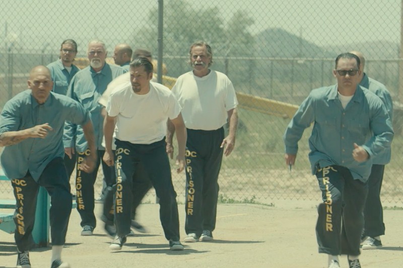 Grupa mężczyzn ubranych w niebieskie spodnie z napisem WIĘZIEŃ idzie i biega na zewnątrz, po dziedzińcu więziennym. W tle widać siatkę ogrodzeniową, góry i suchy krajobraz.