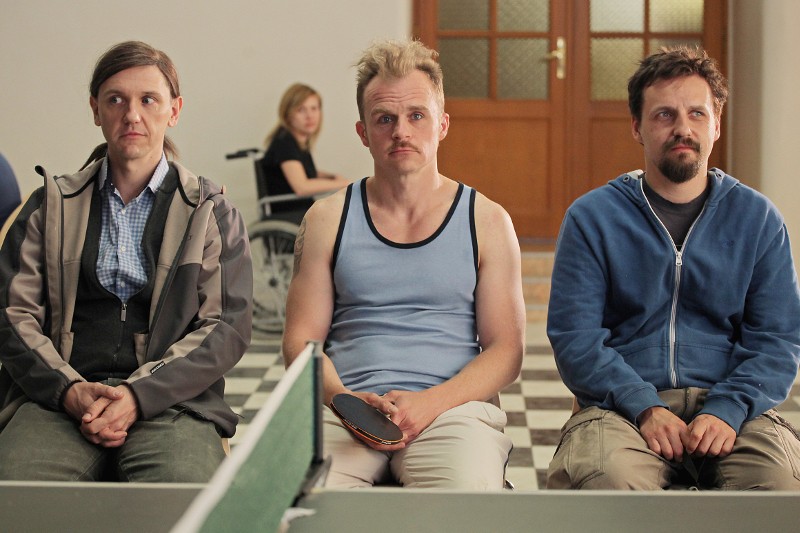 Trzy osoby siedzą obok siebie na ławce w pomieszczeniu, patrząc przed siebie. Osoba pośrodku trzyma paletkę do ping-ponga. W tle widać kobietę na wózku inwalidzkim, a za nimi drzwi z panelami szklanymi.