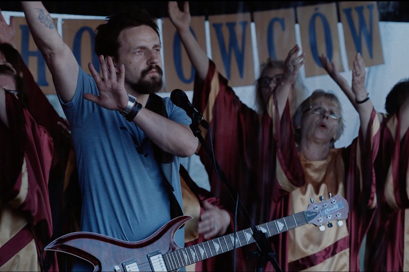 Mężczyzna z gitarą stoi przy mikrofonie z jedną ręką uniesioną do góry, podczas gdy grupa śpiewaków chóru w bordowo-złotych szatach stoi za nim, również podnosząc ręce i śpiewając.