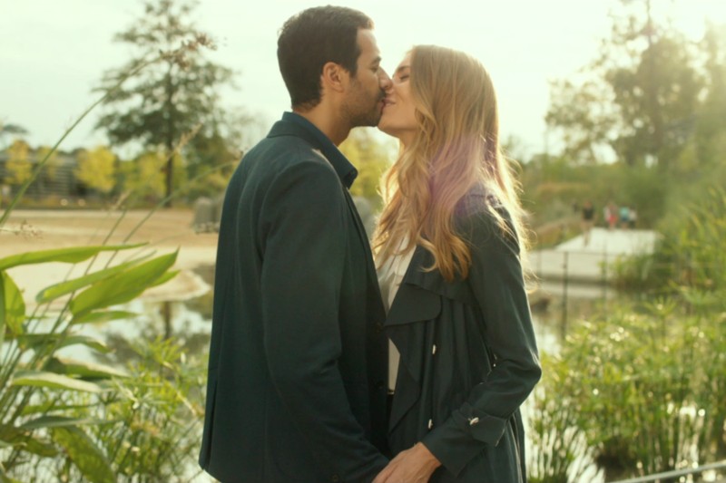 Mężczyzna i kobieta stoją blisko siebie na zewnątrz, w pobliżu stawu, dzieląc się delikatnym pocałunkiem. Światło słoneczne przesącza się przez drzewa za nimi, tworząc ciepłą, romantyczną atmosferę. Oboje mają na sobie ciemne kurtki.