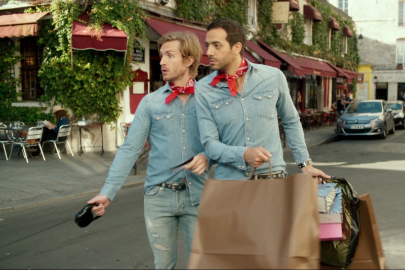 Dwóch mężczyzn ubranych podobnie w dżinsowe koszule i czerwone bandany przechodzi przez ulicę, niosąc torby na zakupy, patrząc w różnych kierunkach. Wyglądają na zaskoczonych lub czujnych. W tle widać kawiarnię na świeżym powietrzu i samochody.