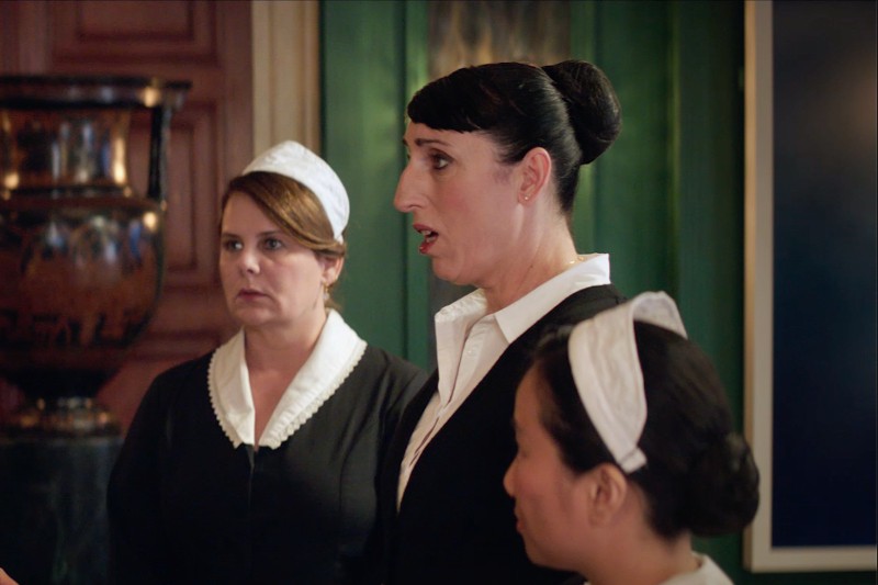 Trzy kobiety ubrane jak pokojówki w czarne mundury z białymi kołnierzykami i nakryciami głowy stoją w pomieszczeniu, sprawiając wrażenie uważnych i poważnych, na tle ciemnozielonej ściany i przedmiotów dekoracyjnych.