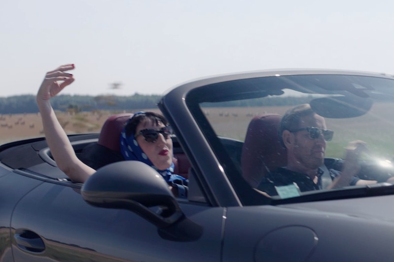 Kobieta ubrana w niebieski szalik i okulary przeciwsłoneczne wypoczywa na siedzeniu pasażera kabrioletu, za kierownicą siedzi mężczyzna; oboje wyglądają na zrelaksowanych, w tle widać otwarte pola.