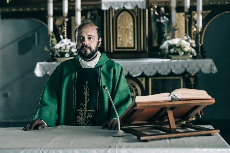Ksiądz w zielonych szatach stoi za ołtarzem w kościele, na drewnianym podium leży otwarta księga, w tle świece i kwiaty.