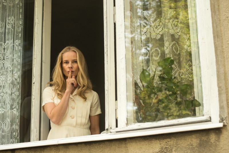 Kobieta w białej sukience stoi przy otwartym oknie, trzymając palec na ustach w geście „shh”. Po obu stronach wiszą koronkowe firanki, a na parapecie stoi zielona roślina.