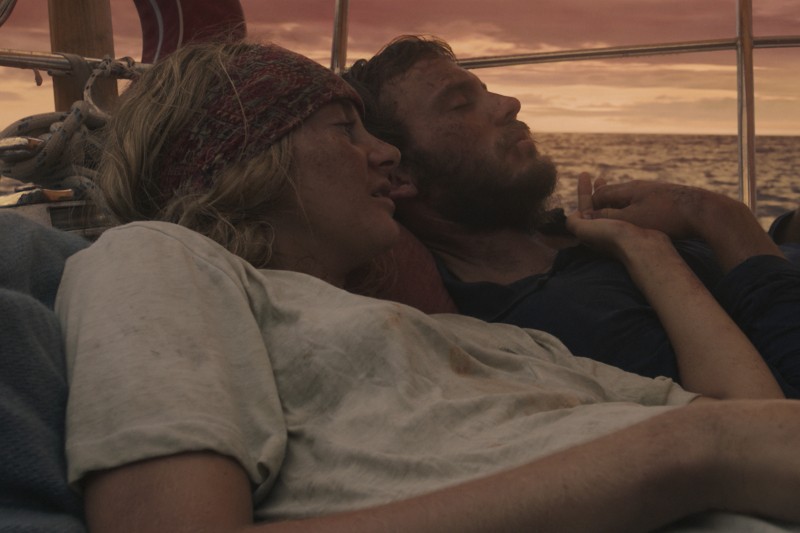 Dwie osoby z potarganymi włosami i brudnymi ubraniami leżą blisko siebie na żaglówce o zachodzie słońca. Wyglądają na wyczerpane, ale pocieszone sobą nawzajem. W tle widać ocean i niebo.