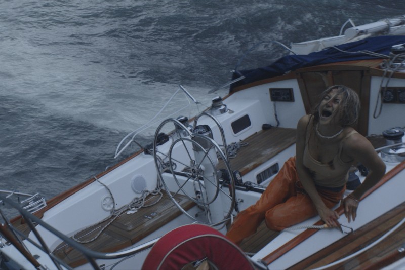 Osoba ubrana w pomarańczowy strój siedzi w rozpaczy na pokładzie żaglówki, trzyma się za bok i krzyczy, a łódź otaczają wzburzone fale oceanu.