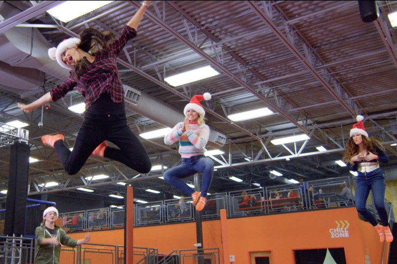 Cztery osoby w czapkach Mikołaja skaczą na trampolinach w krytym parku. Jedna kobieta w kraciastej koszuli jest w powietrzu z uniesionymi rękami. W tle widać pomarańczowe ściany i znak Chill Zone.