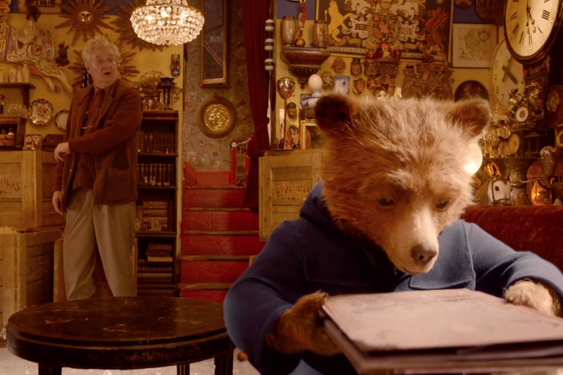Niedźwiedź ubrany w niebieski płaszcz spogląda na książkę w przytulnym, zagraconym antykwariacie, podczas gdy starszy mężczyzna stoi w tle przy schodach wyłożonych czerwonym dywanem, otoczony różnymi zabytkowymi przedmiotami i dekoracjami.