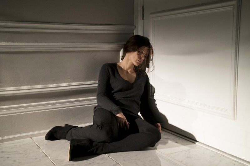 Kobieta ubrana na czarno siedzi na podłodze, oparta o zamknięte białe drzwi w słabo oświetlonym pokoju, z pochyloną głową i zamkniętymi oczami. Wygląda na smutną i wyczerpaną.