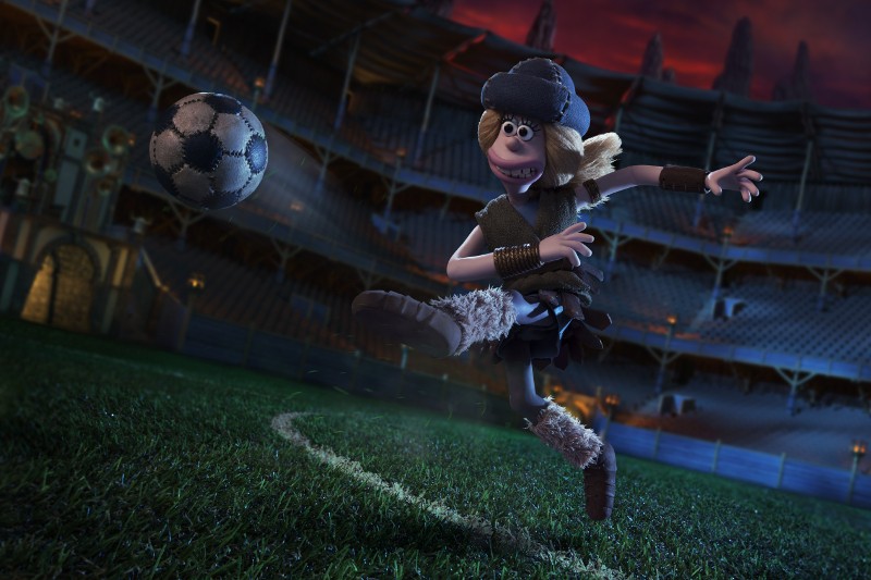 Postać z gliny ubrana w strój w stylu średniowiecznym i hełm przygotowuje się do kopnięcia piłki nożnej na dużym, słabo oświetlonym stadionie.