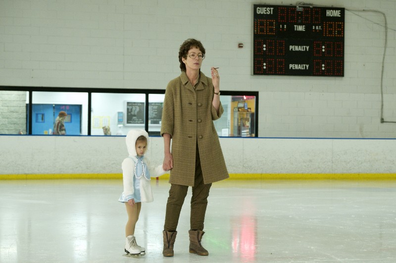 Kobieta w brązowym płaszczu stoi na lodowisku, trzymając za rękę młodą dziewczynę w białym stroju do jazdy na łyżwach. Stoją przed tablicą wyników i obie patrzą w lewą stronę obrazu.