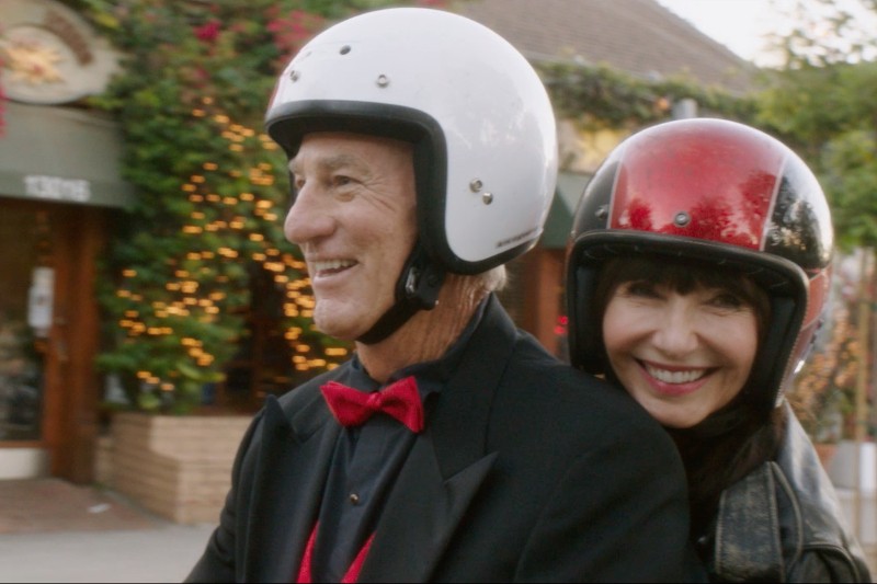 Starszy mężczyzna w białym kasku i czerwonej muszce uśmiecha się, jadąc na skuterze, a kobieta w czerwonym kasku uśmiecha się za nim. Wyglądają na szczęśliwych, w tle widać świąteczne światła i zieleń.