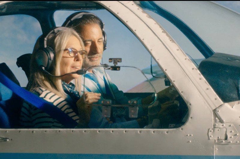 Dwie osoby noszące słuchawki siedzą w kokpicie małego samolotu, uśmiechając się i patrząc przez okno. Promienie słońca przeświecają przez nie, oświetlając wnętrze i ich twarze.