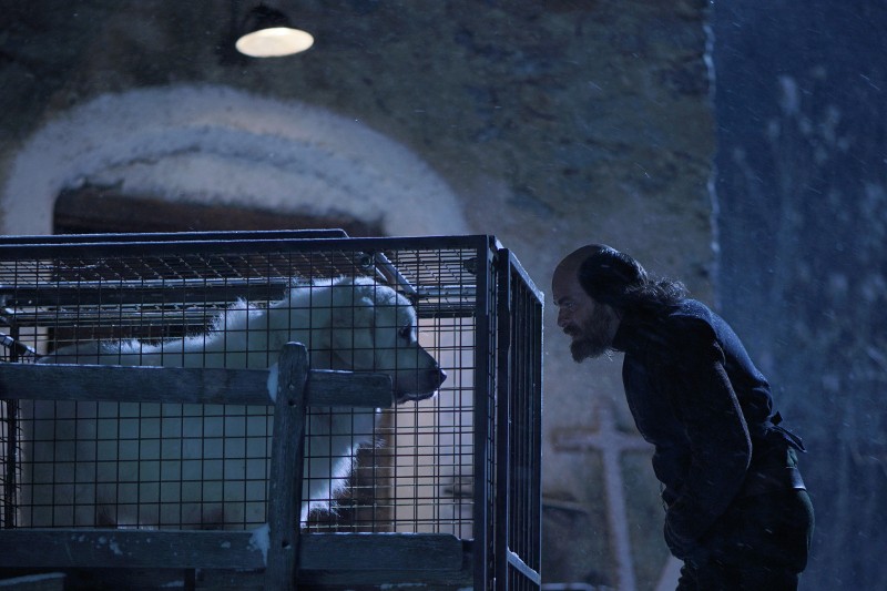 Mężczyzna z długimi włosami i brodą pochyla się nocą w stronę dużego, puszystego, białego psa w klatce. W tle pada śnieg i widać kamienny budynek.