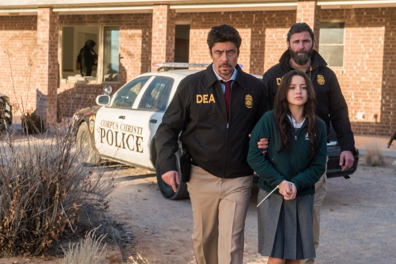 Dwóch mężczyzn w kurtkach policyjnych, jeden z napisem DEA, eskortuje młodą dziewczynę w szkolnym mundurku obok radiowozu przed ceglanym budynkiem. Jeden z funkcjonariuszy trzyma dziewczynę za rękę, gdy idą.