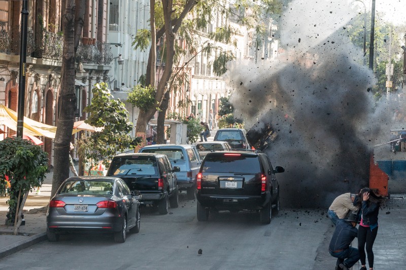 Duża eksplozja wyrzuca dym i gruz w powietrze na miejskiej ulicy, w pobliżu kilku pojazdów i dwóch osób szukających schronienia na chodniku. Drzewa i budynki wyznaczają ruchliwą miejską scenę.