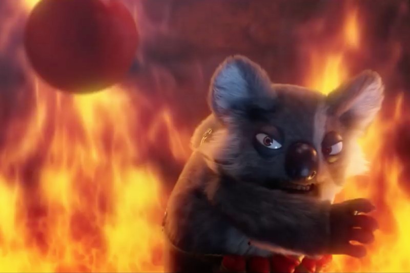 Ożywiony koala o groźnym wyrazie twarzy stoi na tle buchających płomieni, przygotowując się do rzutu czerwoną piłką.