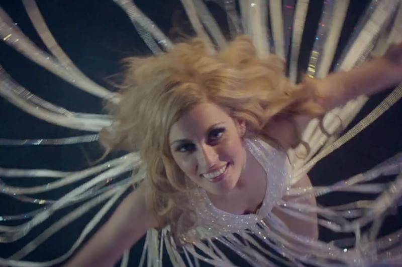 Kobieta o długich, blond włosach uśmiecha się do aparatu, ubrana w błyszczący strój ze srebrnymi wstążkami rozchodzącymi się wokół niej, co tworzy dynamiczny efekt rozbłyskującej gwiazdy.