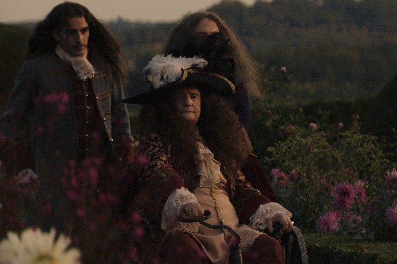 Trzech mężczyzn w wymyślnych strojach z XVII wieku stoi w ogrodzie z kwitnącymi kwiatami. Jeden siedzi na wózku inwalidzkim, ma na sobie duży kapelusz z piórami, podczas gdy pozostali, z długimi włosami, stoją za nim.