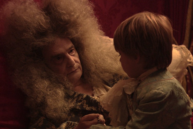 Starsza osoba z dużymi, kręconymi siwymi włosami i ozdobnym ubraniem leży w łóżku, patrząc poważnie na małe dziecko w historycznym stroju, które siedzi obok nich. Tło jest słabo oświetlone i bogato zdobione.