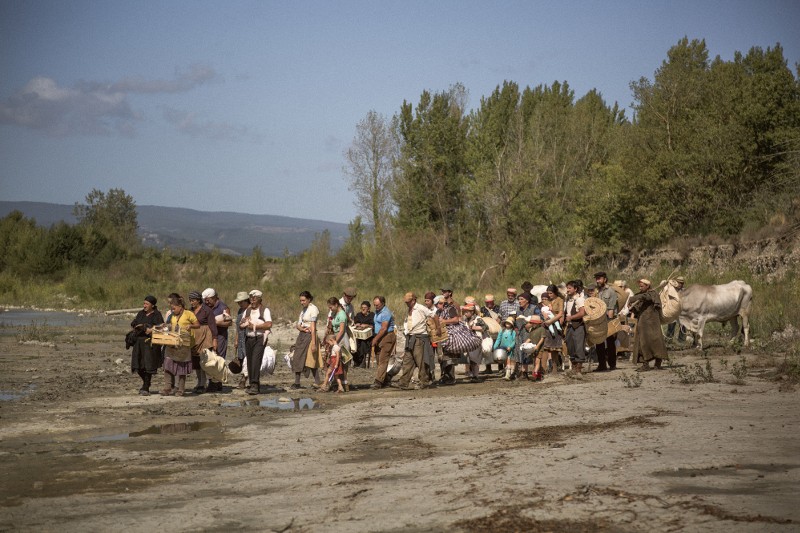 Grupa ludzi, niektórzy niosący tobołki i dzieci, idzie z osłami błotnistą ścieżką wzdłuż rzeki, otoczoną drzewami i zielenią, pod częściowo zachmurzonym niebem.