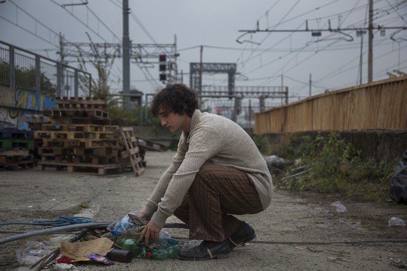 Młody mężczyzna w beżowym swetrze kuca na zewnątrz w pobliżu torów kolejowych, zbierając plastikowe butelki i śmieci z ziemi. Obszar jest usiany gruzem, drewnianymi paletami i otoczony konstrukcjami przemysłowymi i liniami energetycznymi.