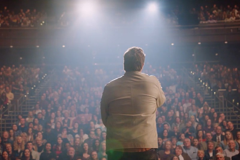 Osoba stoi na scenie naprzeciwko dużej, siedzącej publiczności w teatrze oświetlonym jasnymi światłami, a rzędy ludzi uważnie ją obserwują.