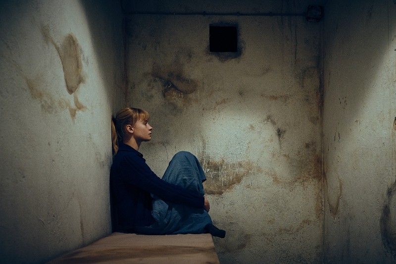 Kobieta siedzi sama na ławce w małej, słabo oświetlonej, betonowej celi o poplamionych ścianach, obejmuje kolana i patrzy przed siebie zamyślonym wzrokiem.