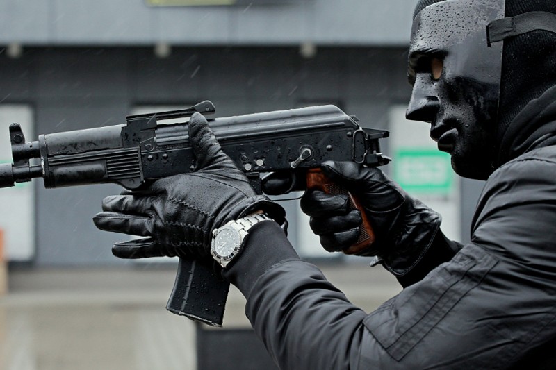 Osoba ubrana w czarną maskę, rękawice i zegarek celuje z czarnego karabinu szturmowego. Tło jest rozmyte, skupiając uwagę na uzbrojonej osobie w ciemnym ubraniu.