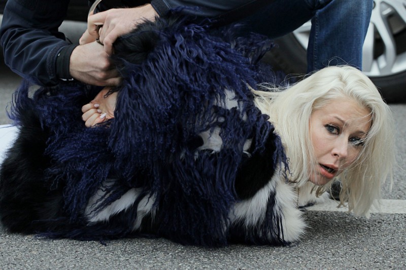 Kobieta z długimi blond włosami ubrana w czarno-biały płaszcz leży na ziemi, podczas gdy ktoś trzyma jej ręce za plecami, najwyraźniej ją powstrzymując. Wygląda na zmartwioną i leży na utwardzonej nawierzchni w pobliżu pojazdu.