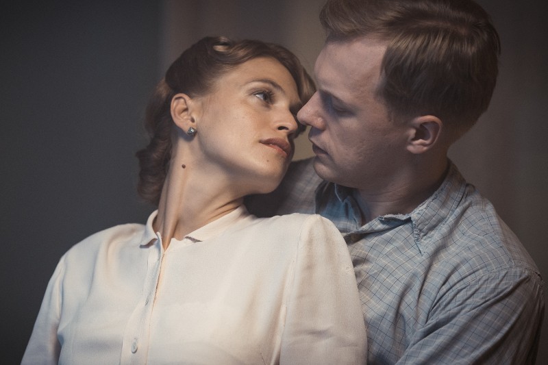 Kobieta w białej bluzce odchyla się do tyłu i spogląda na mężczyznę w kraciastej koszuli, który ją obejmuje. Oboje przeżywają intensywną, intymną chwilę w delikatnym oświetleniu.