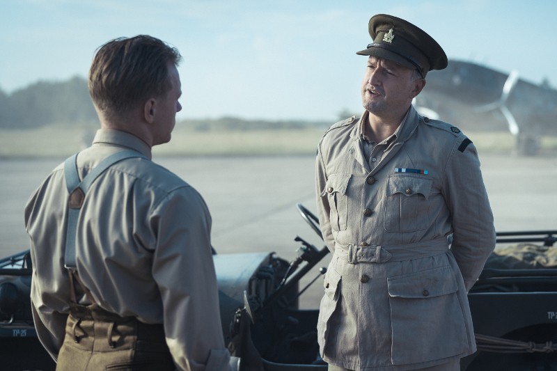 Dwóch mężczyzn w mundurach wojskowych stoi i rozmawia na zewnątrz, w pobliżu jeepa, z samolotem i otwartym polem w tle. Jeden mężczyzna stoi twarzą do kamery, a drugi jest odwrócony plecami.