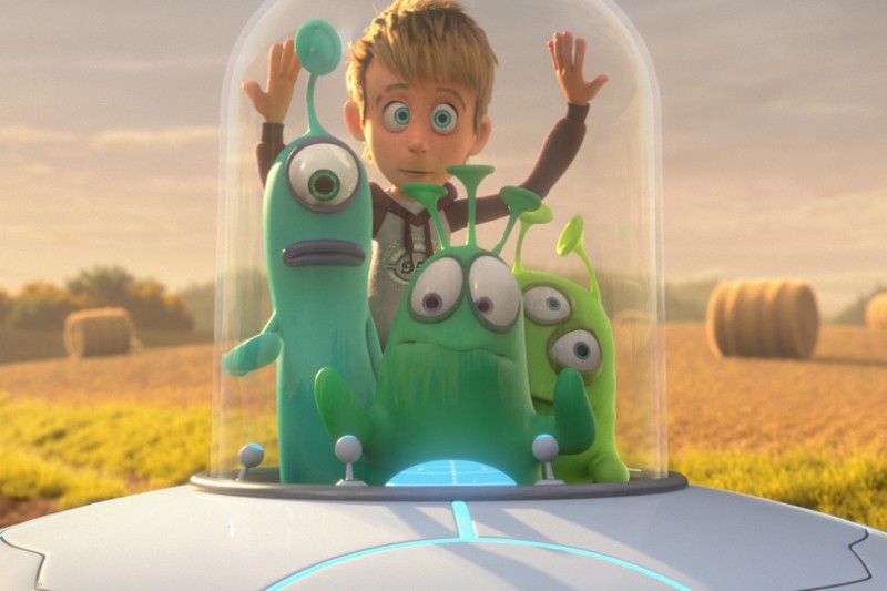 Młody chłopiec o blond włosach stoi wewnątrz szklanej kopuły z trzema kolorowymi, kreskówkowymi przybyszami na statku kosmicznym, a całość rozgrywa się na polu z belami siana w tle.