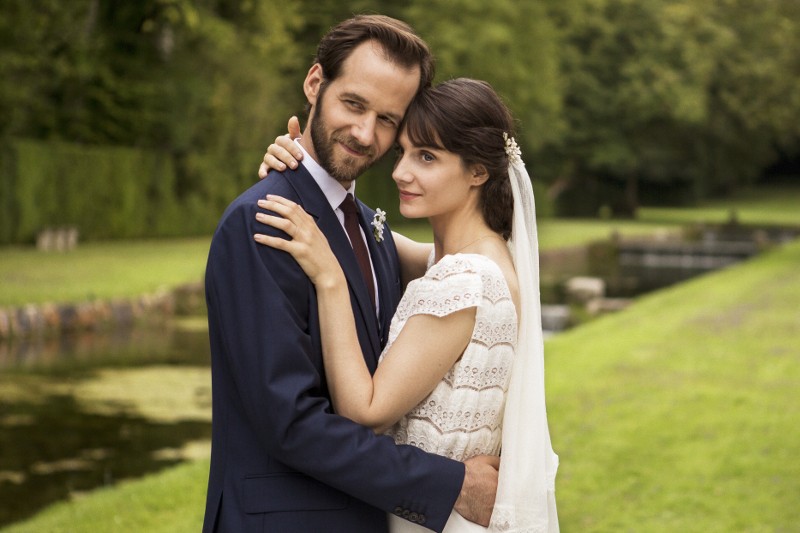 Panna młoda i pan młody obejmują się na zewnątrz, uśmiechając się do kamery. Panna młoda ma na sobie białą suknię i welon, a pan młody ciemny garnitur. Stoją na zielonej trawie z drzewami i kanałem wodnym w tle.