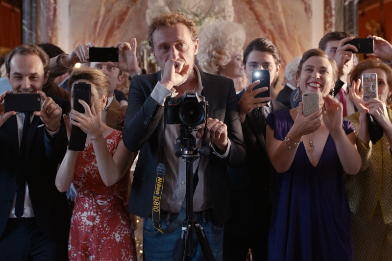 Grupa ludzi, niektórzy uśmiechnięci, stoją blisko siebie, robiąc zdjęcia i filmy smartfonami i aparatami na wydarzeniu, prawdopodobnie weselu lub uroczystości. Mężczyzna z aparatem na statywie jest w centrum.