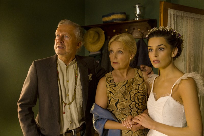 Trzy osoby stoją razem w słabo oświetlonym pokoju, wyglądając na zaskoczone. Starszy mężczyzna w garniturze, kobieta w średnim wieku w złotej sukience i młoda kobieta w białej sukni ślubnej i kwiatowym nakryciu głowy trzymają się za ręce.