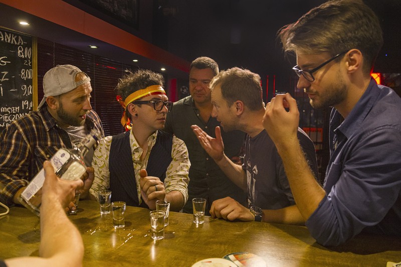 Pięciu mężczyzn zebrało się przy barze z kieliszkami przed sobą; jeden nalewa klarownego drinka, podczas gdy inni rozmawiają ożywionym tonem. Jeden mężczyzna nosi opaskę na głowie i okulary, a atmosfera jest ożywiona.