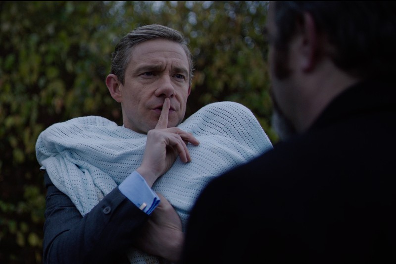 Mężczyzna trzyma na rękach dziecko owinięte w niebieski kocyk i gestem ręki przystawia palec do ust, nakazując ciszę, stojąc twarzą do innej osoby na zewnątrz.